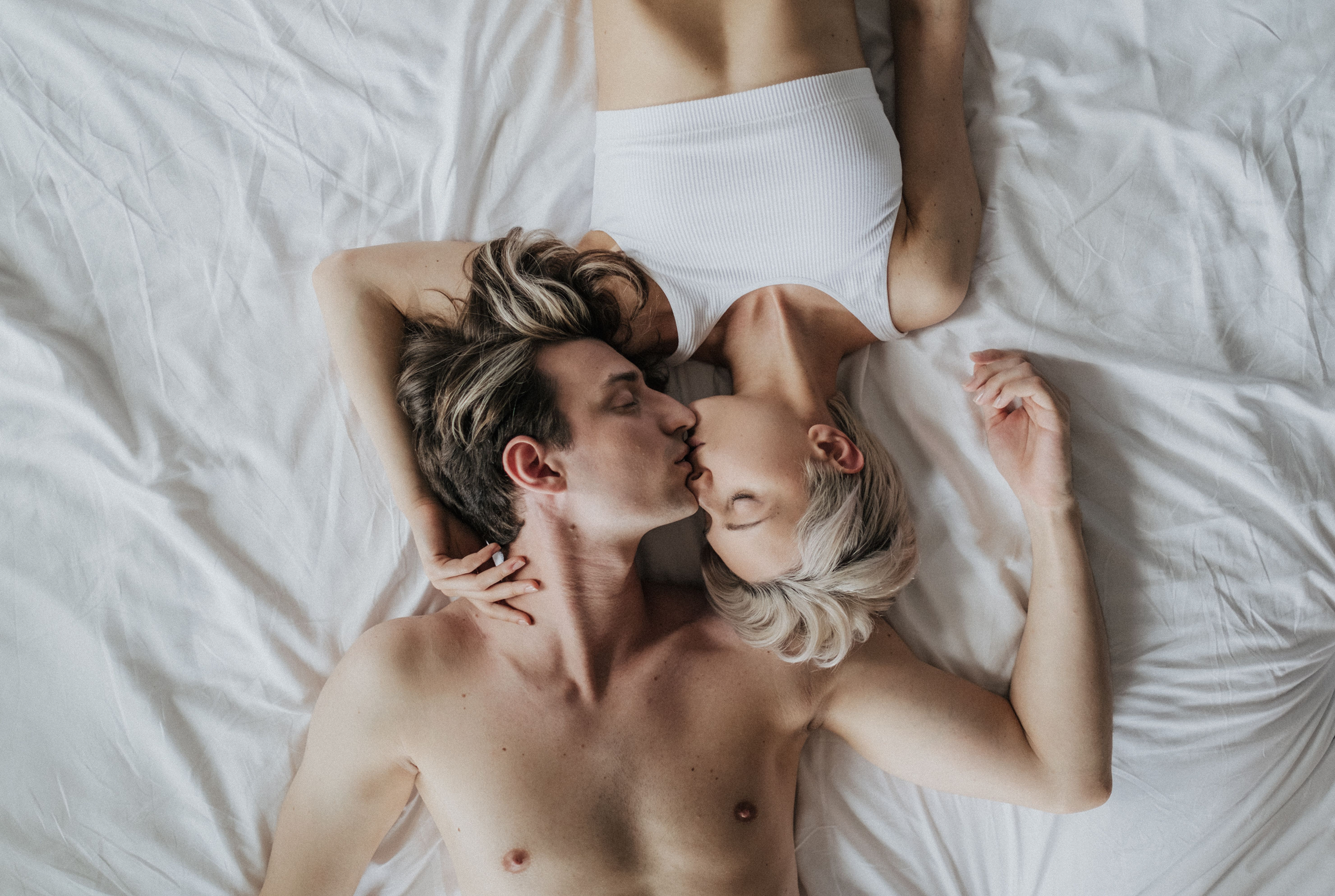 A man and a woman lying on white bedsheets with their heads together, leaning to kiss